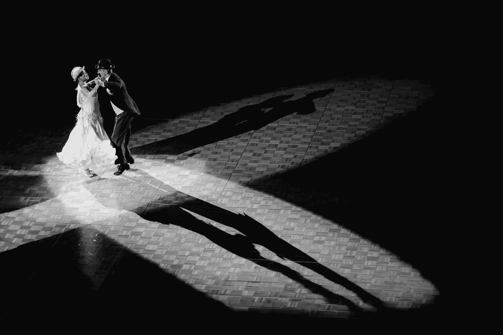 A couple in formal dress dancing a ballroom waltz under a single spotlight on a wooden floor, representative of the waltz danced at the Strauss Dinner Show